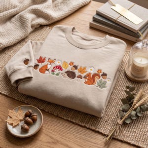 Könnte beinhalten: Ein beigefarbenes Sweatshirt mit Rundhalsausschnitt und einem horizontalen Stickmuster aus Waldmotiven, darunter ein Fuchs, ein Igel, ein Eichhörnchen, rote und braune Pilze, Herbstblätter, Eicheln und kleine weiße Blumen am Ärmel.