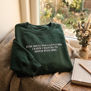 以下が含まれることがあります： 深いフォレストグリーンのクルーネックトレーナー。白い刺繍で「EVER SINCE I WAS A LITTLE GIRL I KNEW I WANTED TO ARGUE WITH MEN」という文字が施され、袖口と襟元はリブ編みになっています。質感のあるベージュの布地の上に置かれています。
