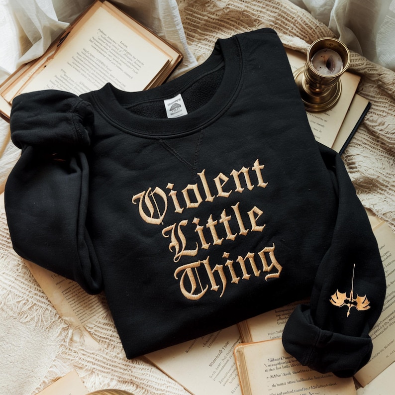 May include: Black sweatshirt with the embroidered text "Violent Little Thing" in a vintage font. The sweatshirt has a crew neck and long sleeves. A small gold-colored emblem is on the left sleeve. The sweatshirt is displayed on a stack of old books.