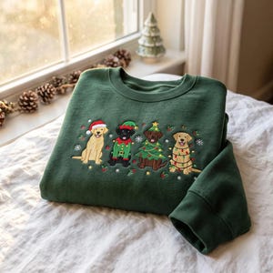 Puede incluir: Una sudadera de cuello redondo verde oscuro con un diseño bordado de cuatro labradores. Los perros llevan ropa festiva, incluyendo un gorro de Papá Noel, un disfraz de elfo, un traje de árbol de Navidad y una guirnalda de luces de colores.