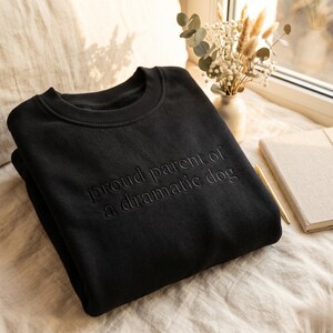 May include: Black crewneck sweatshirt with the embroidered text "proud parent of a dramatic dog." The sweatshirt is folded on a neutral-toned surface, with a vase of dried flowers and a notebook in the background.