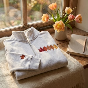 May include: A light gray heathered quarter-zip sweatshirt featuring embroidered autumn leaves in shades of red, orange, and yellow on the chest and a single matching leaf embroidered on the left sleeve cuff, with a silver-toned metal zipper.