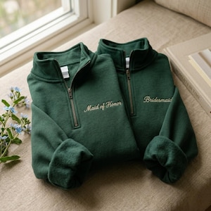 Op de afbeelding: Twee donkergroene sweatshirts met een kwartrits. Eén sweatshirt heeft "Maid of Honor" geborduurd in een sierlijk lettertype, de andere zegt "Bridesmaid". Beide hebben een rits en opgerolde manchetten. De sweatshirts liggen op een neutrale ondergrond.