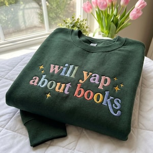 May include: A dark green sweatshirt featuring the embroidered phrase "will yap about books" in pastel shades. The garment is folded, showing the sleeves. The design includes star motifs.