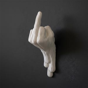 Può includere: Una scultura da parete bianco opaco a forma di mano che fa il gesto del dito medio. Il design presenta una superficie liscia e solida con un effetto gocciolante alla base, creando un orientamento verticale che si estende verso il basso.