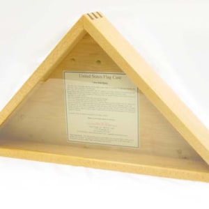 May include: A wooden triangle-shaped flag case with a clear glass front. The case is made of light wood and has a dark wood trim. The text "United States Flag Case" is printed on the glass.