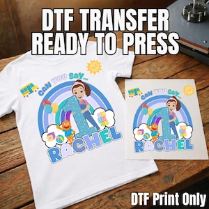 May include: White t-shirt with a colourful graphic featuring a girl, the number 1, and the name "RACHEL". The design includes a rainbow, sun, and the text "CAN YOU SAY..." and "DTF TRANSFER READY TO PRESS".