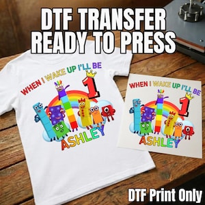 May include: A white t-shirt and a square DTF transfer sheet featuring a colourful graphic of cartoon number blocks, a rainbow, a large number one with a crown, and the text "WHEN I WAKE UP I'LL BE 1" and the name "ASHLEY" in rainbow-coloured letters.