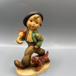 May include: A ceramic figurine of a young boy in a green jacket, brown pants, and a black hat, holding a red umbrella. A small brown dog walks beside him. The figurine is on a light-colored oval base.