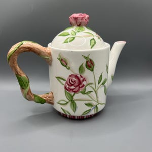 May include: A white ceramic teapot with a floral design featuring pink roses and green leaves. The teapot has a twisted brown handle and a lid with a pink rose on top.