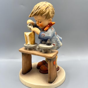 May include: A ceramic figurine of a young boy in a blue outfit with red accents, working at a pottery wheel. The figurine is on a small wooden table with brown shoes at the base. The boy has blonde hair and is focused on his work.