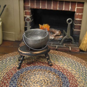 May include: Miniature metal cauldron with a handle, resting on a small, dark wooden stool. The scene includes a brick fireplace, a broom, and a colorful braided rug, creating a rustic, cozy atmosphere. The cauldron is approximately 2 inches in diameter.
