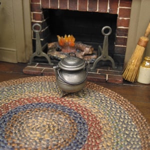 May include: Miniature metal pot with a lid and handle, placed on a colorful braided rug. The rug has concentric circles of red, blue, and tan. A brick fireplace with a burning fire is in the background, along with a broom and other rustic accessories.