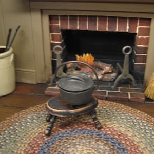May include: A miniature scene featuring a fireplace with a brick surround, a black pot on a small wooden stool, and a braided rug. A broom and a ceramic jug are placed nearby. A ceramic crock with tools is on the left.