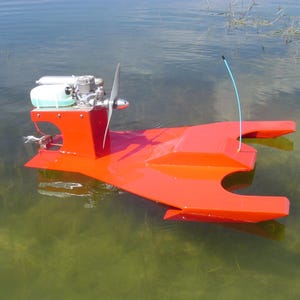 Könnte beinhalten: Ein leuchtend rotes Modellboot mit Propeller und Motor schwimmt auf dem Wasser. Das Boot hat ein stromlinienförmiges Design mit einem zentralen Körper und zwei seitlichen Pontons. Eine blaue Antenne erstreckt sich nach oben.