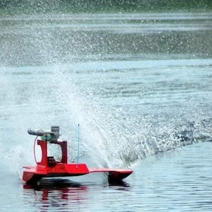 May include: A red remote-controlled boat speeding across a body of water, creating a large splash. The boat has a small engine and a tall antenna. The water is a light blue color, and the background is a blurry green.