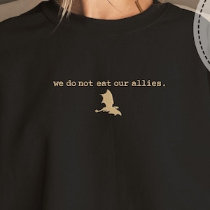 Puede incluir: Sudadera negra con un drag&oacute;n bordado en oro y el texto "we do not eat our allies."