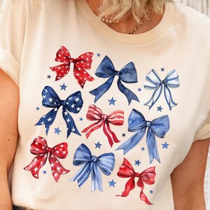 May include: A cream-coloured t-shirt with a watercolour design of red, white, and blue bows and stars. The bows feature various patterns, including stripes and stars, and are spread across the shirt.