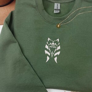 May include: A dark green sweatshirt with a white embroidered design of a stylized creature with a symbol on its chest. The sweatshirt has a gold chain necklace with a small gold pendant.