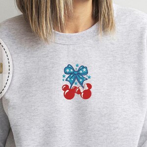 Op de afbeelding: Een lichtgrijs sweatshirt met een patriottisch ontwerp. Het ontwerp toont een blauwe strik met witte sterren en rode Mickey Mouse-vormige kersen. De sweatshirt is een comfortabele, casual stijl.