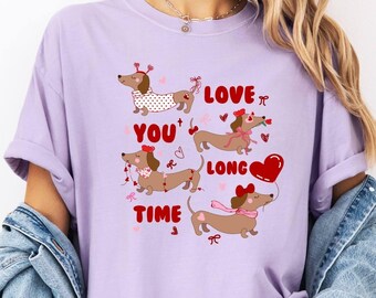 May include: A lilac t-shirt with a graphic of dachshund dogs, hearts, and bows. The text "LOVE YOU LONG TIME" is above the dogs. The shirt has short sleeves and a relaxed fit. The image is of a casual style.