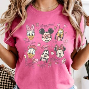 May include: A pink t-shirt featuring a collage of Disney characters, including Mickey Mouse, Minnie Mouse, Donald Duck, and Daisy Duck. The design includes hearts and the text "Mickey Mouse".