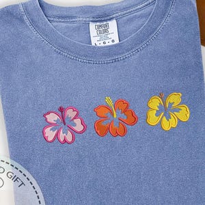 May include: A blue t-shirt with three embroidered hibiscus flowers in pink, orange, and yellow. The shirt has a crew neck and a tag that reads "Comfort Colors". The flowers are detailed with contrasting outlines.