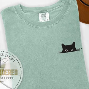 K&ouml;nnte beinhalten: Salbeigr&uuml;nes Comfort Colors T-Shirt mit einem schwarzen Katzenmotiv, das auf der Vorderseite aufgestickt ist. Die Katze schaut &uuml;ber den Rand des Shirts. Das T-Shirt hat einen Rundhalsausschnitt und kurze &Auml;rmel. Das Etikett lautet "L - G - G".