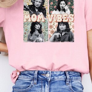 Pu&ograve; includere: Maglietta rosa con un collage di quattro volti di donne e motivi floreali. Il testo "MOM VIBES" &egrave; stampato in un font retr&ograve;.