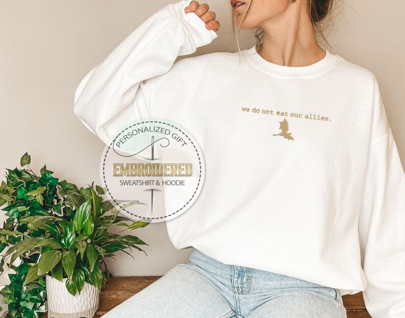 Puede incluir: Sudadera blanca con el texto "we do not eat our allies." y un peque&ntilde;o dise&ntilde;o de drag&oacute;n bordado. La sudadera es un regalo personalizado.