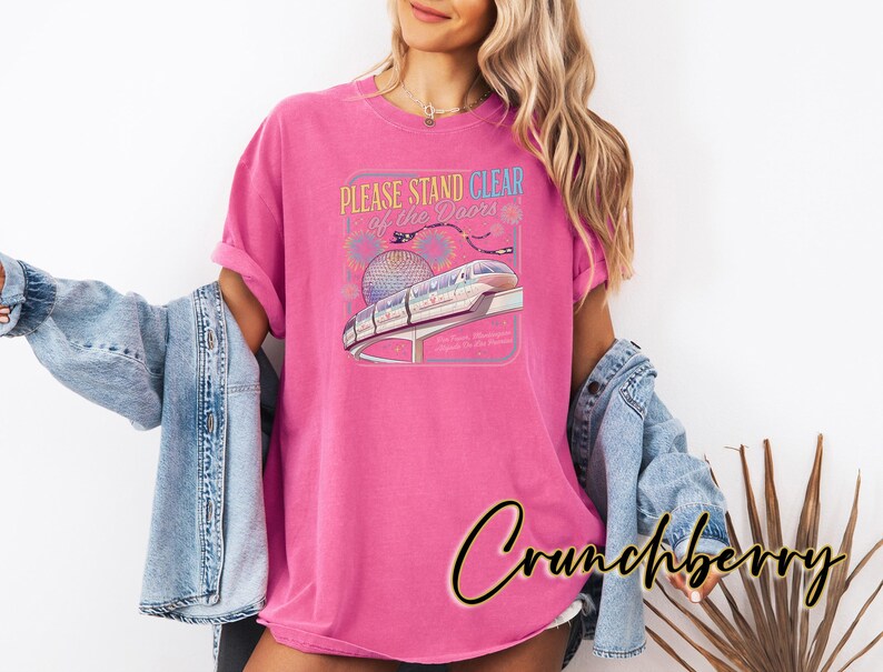 May include: A magenta t-shirt with a graphic featuring a monorail, the text "PLEASE STAND CLEAR of the DOORS", and a stylized depiction of a sphere. The shirt is worn with a light blue denim jacket.