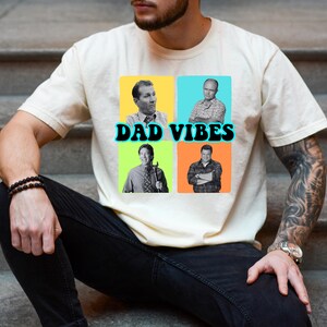 May include: Cream-colored t-shirt featuring a graphic with four images of men in colorful squares. The text "DAD VIBES" is in black with a blue outline. The shirt is worn by a person sitting on concrete steps.