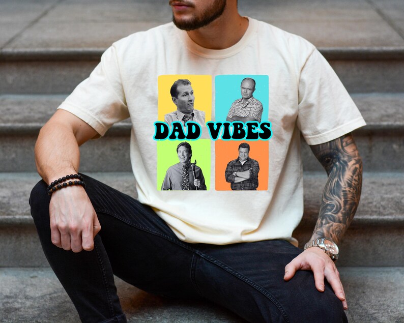 May include: Cream-colored t-shirt featuring a graphic with four images of men in colorful squares. The text "DAD VIBES" is in black with a blue outline. The shirt is worn by a person sitting on concrete steps.