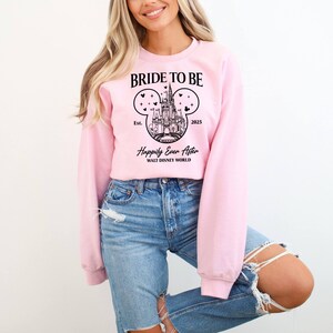 Puede incluir: Sudadera rosa con un gr&aacute;fico negro del castillo de Cenicienta en forma de oreja de Mickey Mouse. El texto dice "Bride to Be Est. 2023 Happily Ever After Walt Disney World".