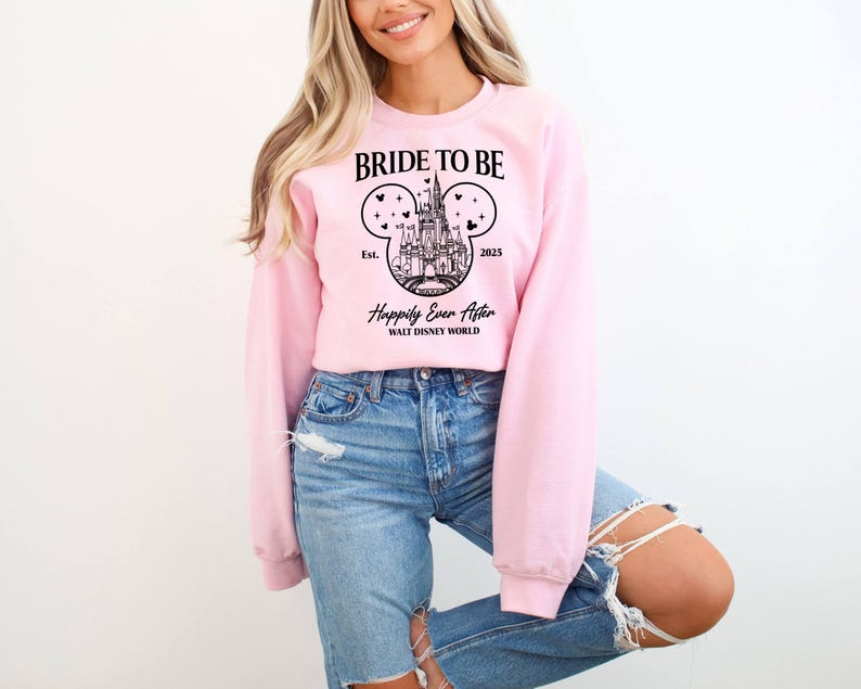 Puede incluir: Sudadera rosa con un gr&aacute;fico negro del castillo de Cenicienta en forma de oreja de Mickey Mouse. El texto dice "Bride to Be Est. 2023 Happily Ever After Walt Disney World".