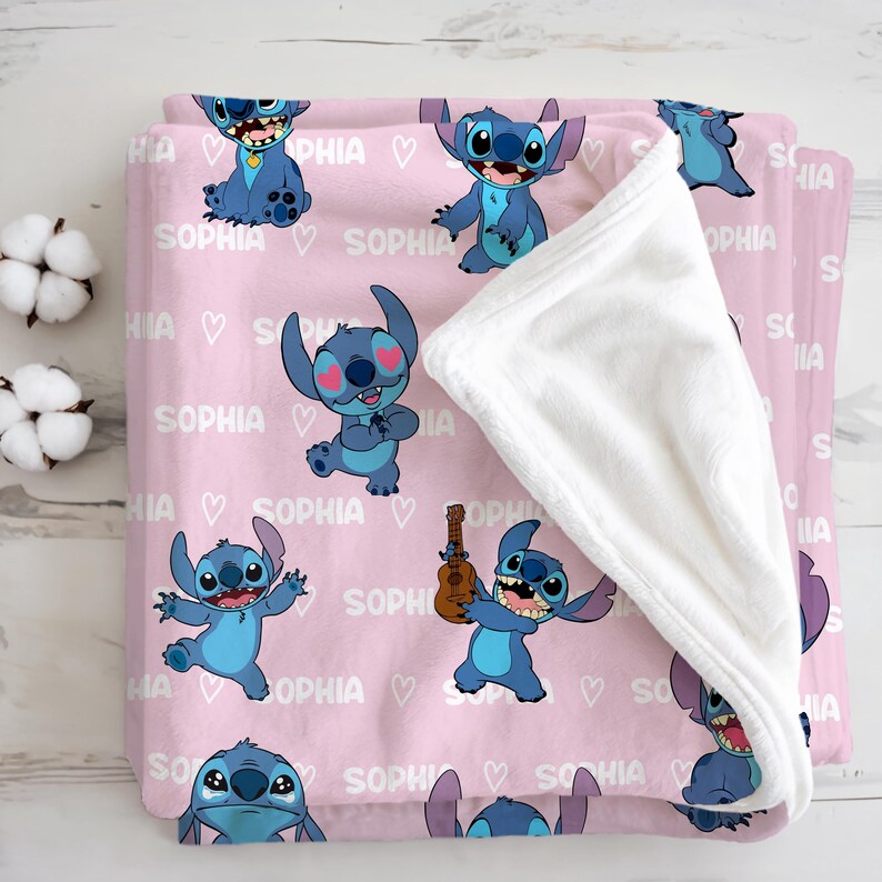 May include: A pink fleece blanket featuring cartoon character Stitch in various poses, with the name "Sophia" and hearts printed across the surface. The blanket has a white, fluffy underside.