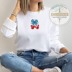 Op de afbeelding: Witte sweatshirt met een geborduurd ontwerp met een blauwe strik, rode sterren en rode bollen. De sweatshirt heeft lange mouwen en een ronde hals. De tekst "PERSONALIZED GIFT EMBROIDERED SWEATSHIRT & HOODIE" is ook zichtbaar.