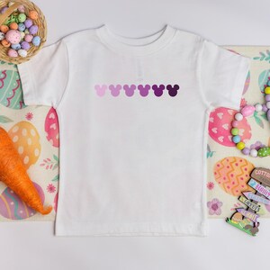 Puede incluir: Camiseta blanca con una fila de cabezas de Mickey Mouse en degradado de morado en la parte delantera. La camiseta est&aacute; sobre una mesa con un patr&oacute;n de huevos de Pascua coloridos.