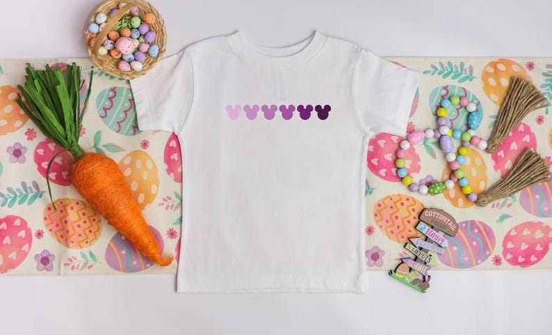 Puede incluir: Camiseta blanca con una fila de cabezas de Mickey Mouse en degradado de morado en la parte delantera. La camiseta est&aacute; sobre una mesa con un patr&oacute;n de huevos de Pascua coloridos.