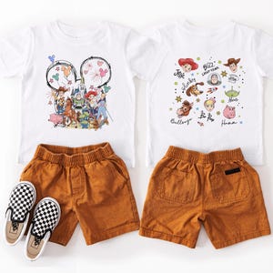 May include: Two white t-shirts with cartoon character graphics, paired with orange shorts and black and white checkered slip-on shoes. The shirts feature characters from a popular animated movie.