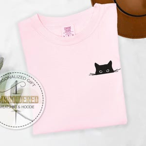 K&ouml;nnte beinhalten: Hellrosa T-Shirt mit einem schwarzen Katzenmotiv. Der Kopf und die Augen der Katze sind &uuml;ber einer Linie zu sehen. Das T-Shirt hat einen Rundhalsausschnitt und ein kleines Etikett. Ein brauner Hut und ein Geschenkanh&auml;nger sind ebenfalls im Bild.