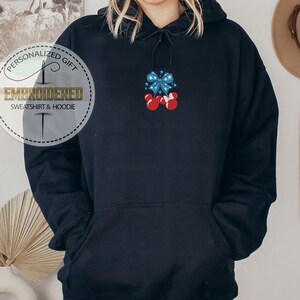 Op de afbeelding: Zwarte hoodie met een voorzak en een capuchon met trekkoord. De hoodie heeft een geborduurd ontwerp van een blauwe strik met rode Mickey Mouse-stijl handschoenen. De tekst "PERSONALIZED GIFT EMBROIDERED SWEATSHIRT & HOODIE" is gedrukt op een rond label.