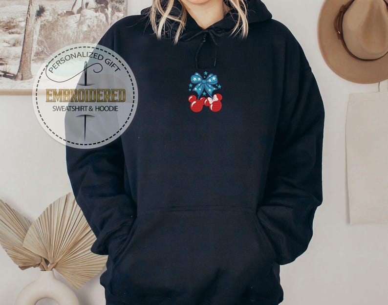 Op de afbeelding: Zwarte hoodie met een voorzak en een capuchon met trekkoord. De hoodie heeft een geborduurd ontwerp van een blauwe strik met rode Mickey Mouse-stijl handschoenen. De tekst "PERSONALIZED GIFT EMBROIDERED SWEATSHIRT & HOODIE" is gedrukt op een rond label.
