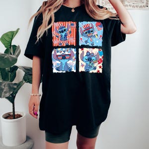 May include: Black t-shirt with a four-panel graphic of a blue cartoon character in patriotic outfits. Each panel shows the character with American flag elements, such as a hat and sunglasses. The design is set against red, white, and blue backgrounds.
