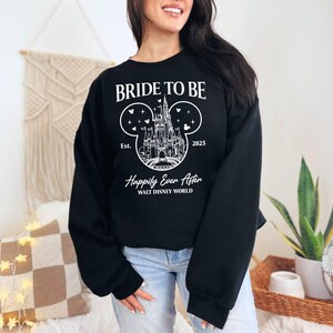 Puede incluir: Sudadera negra con texto blanco que dice "Bride to Be" y un gr&aacute;fico del castillo de Cenicienta dentro de una oreja de Mickey Mouse. El texto "Est. 2025" y "Happily Ever After Walt Disney World" tambi&eacute;n est&aacute;n incluidos.