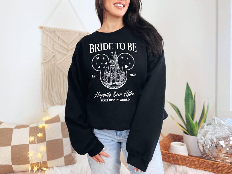 Puede incluir: Sudadera negra con texto blanco que dice "Bride to Be" y un gr&aacute;fico del castillo de Cenicienta dentro de una oreja de Mickey Mouse. El texto "Est. 2025" y "Happily Ever After Walt Disney World" tambi&eacute;n est&aacute;n incluidos.