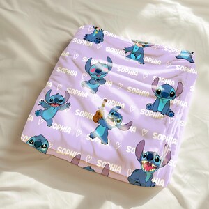 May include: A lavender blanket featuring multiple illustrations of the character Stitch in various poses. The word "SOPHIA" and hearts are printed across the blanket in white. The blanket is folded on a white surface.