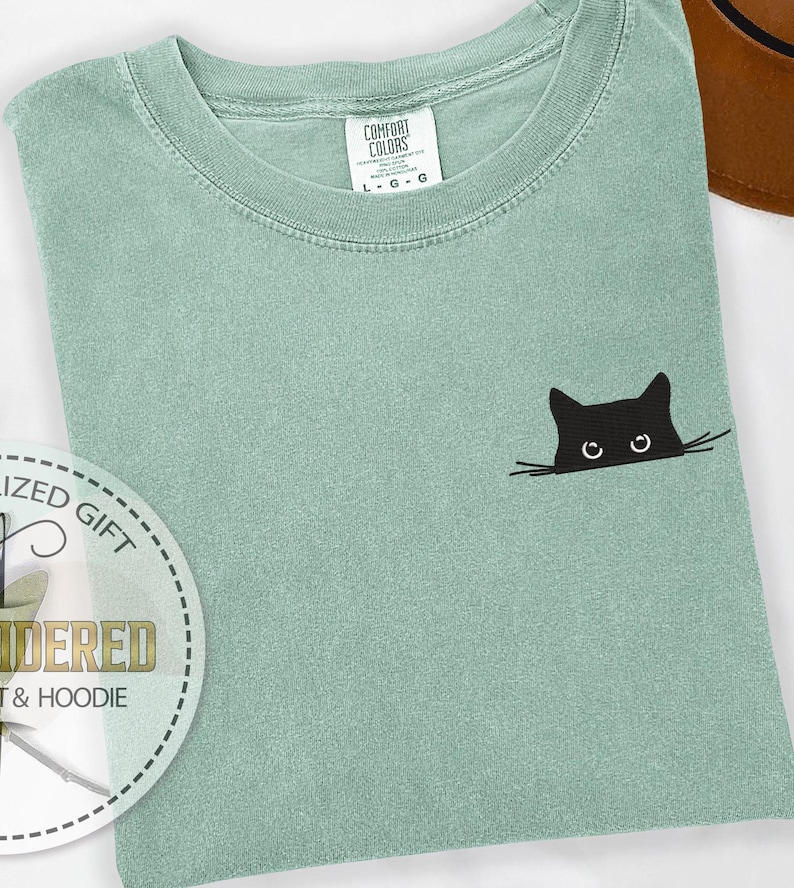 K&ouml;nnte beinhalten: Salbeigr&uuml;nes Comfort Colors T-Shirt mit einem schwarzen Katzenmotiv, das auf der Vorderseite aufgestickt ist. Die Katze schaut &uuml;ber den Rand des Shirts. Das T-Shirt hat einen Rundhalsausschnitt und kurze &Auml;rmel. Das Etikett lautet "L - G - G".