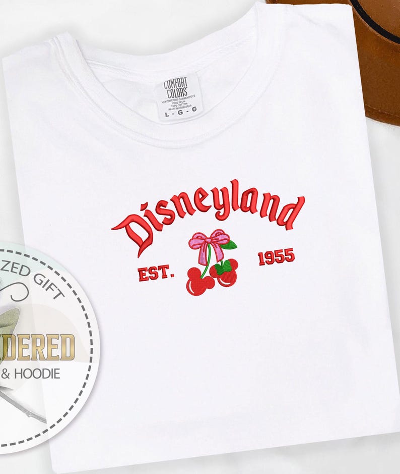 Pu&ograve; includere: Maglietta bianca con un logo Disneyland rosso e una grafica di ciliegie. Il logo dice "Disneyland" con "EST. 1955" sotto.
