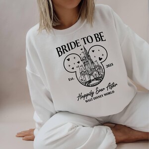 Puede incluir: Sudadera blanca con texto negro que dice "Bride to Be" y un contorno negro del castillo de Cenicienta dentro de una oreja de Mickey Mouse. El texto "Est. 2025 Happily Ever After Walt Disney World" tambi&eacute;n est&aacute; impreso en la sudadera.